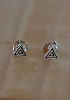 A pair of sterling silver triangular studs  with a rippled texture design, displayed against a wooden background 