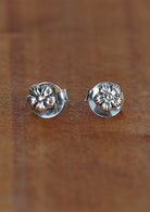 A pair of dainty flower silver studs with delicate bloom design, displayed on a wooden surface