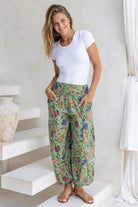 Woman wearing a white rayon top and moss green based floral pants with hands in deep pockets.
