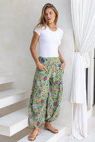 Woman wearing a white top and moss green floral pants with wide elastic waist and ankles standing in front of a white wall 