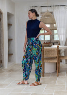 A woman standing side on wearing high-waisted, loose-fit cotton pants with a vibrant multicolor floral print in shades of blue, green, yellow, and pink, paired with a navy blue sleeveless top and brown sandals. 