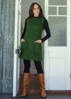Woman wearing a green cotton corduroy sleeveless tunic layered over a black top and leggings and tan boots, with hands in pockets
