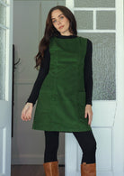Woman wearing a green cotton corduroy sleeveless tunic layered over a black top and leggings and tan boots
