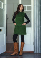 Woman wearing a green cotton corduroy sleeveless tunic layered over a black top and leggings and tan boots, with hands in front pockets