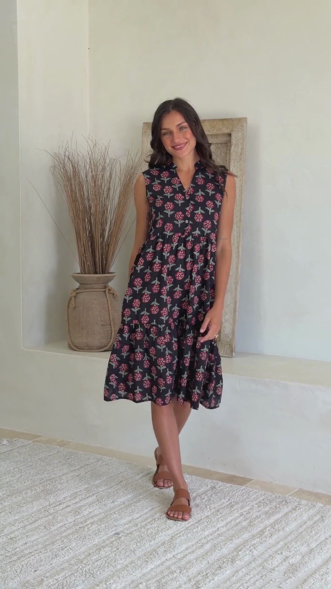 Video of woman wearing a knee length cotton sleeveless dress with buttoned bodice and  two tiered skirt in black based floral print.