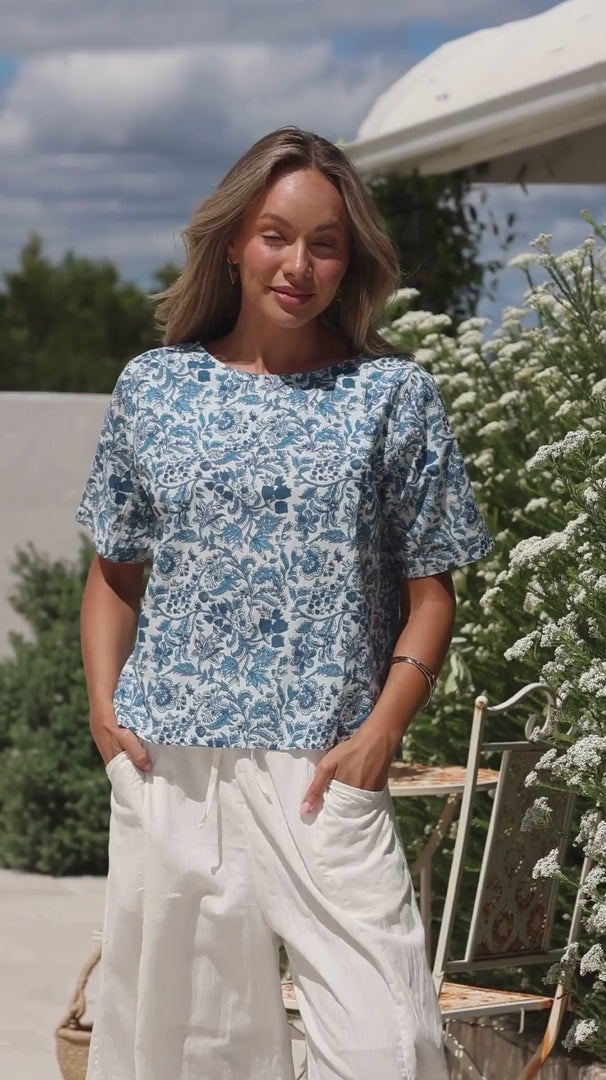Video of a woman wearing a white based blue floral printed relaxed fit cotton top with short sleeves and high neckline
