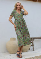 Woman wearing relaxed fit button through dress in moss green based multicoloured floral print with V neckline, pockets and short sleeves.