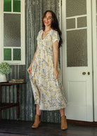 Woman in a white floral cotton maxi dress with full skirt  standing on a verandah of a rustic cottage