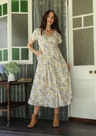 Woman in a white floral cotton dress standing on a verandah of a rustic cottage with her hand in a pocket 