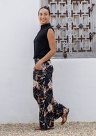 Woman wearing a black sleeveless top and floral cotton pants standing against a white wall with a decorative metal gate.