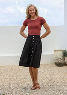 Woman posing in knee length black cotton button-up skirt with belt loops, pockets and box pleats