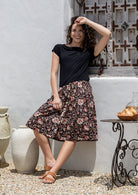Woman leans on wall in a A line, button through 100% cotton skirt with belt loops, box pleats and elastic back waist.