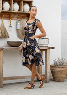 Model wearing with one hand in pocket of a black based floral print dress with scoop neck, cap sleeves and piped detailing at the waist and pockets.