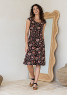 Model standing in front of white wall wearing a retro style 100 percent cotton dress with rust, tan and sand tones on a black base