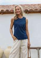 Woman wearing a blue sleeveless cotton top with side bust darts and off white pants standing against a white wall with a terracotta roof.