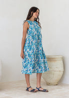 Woman stands side on in cotton sleeveless three tiered midi length dress in aqua and royal blue daisy print