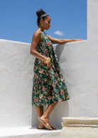 Woman stands in side pose wearing a tiered sleeveless sundress with pockets, high round neck and empire waist.