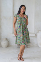 Woman wearing a green floral above knee cotton dress with cap sleeves, standing in a minimalistic room with white walls.