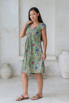 Woman wearing a green floral A-line dress with fitted bodice, cap sleeves and round neckline, standing in a room with white walls and a vase.