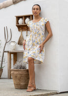 Woman wearing a 100% cotton neutral based floral dress standing against a white wall with wooden shelves.