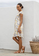 Woman standing sideways wearing a neutral based floral print cotton patterned sundress standing against a plant in the background