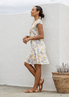Woman in a floral dress standing against a white wall with a plant in the foreground.