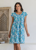 Woman wearing cap sleeved cotton above knee dress in blue and aqua daisy print, with round neckline and pin-tucks across bodice
