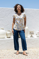 Woman wearing 100% cotton top in neutral based floral print with plain blue pants standing in front of white vases.