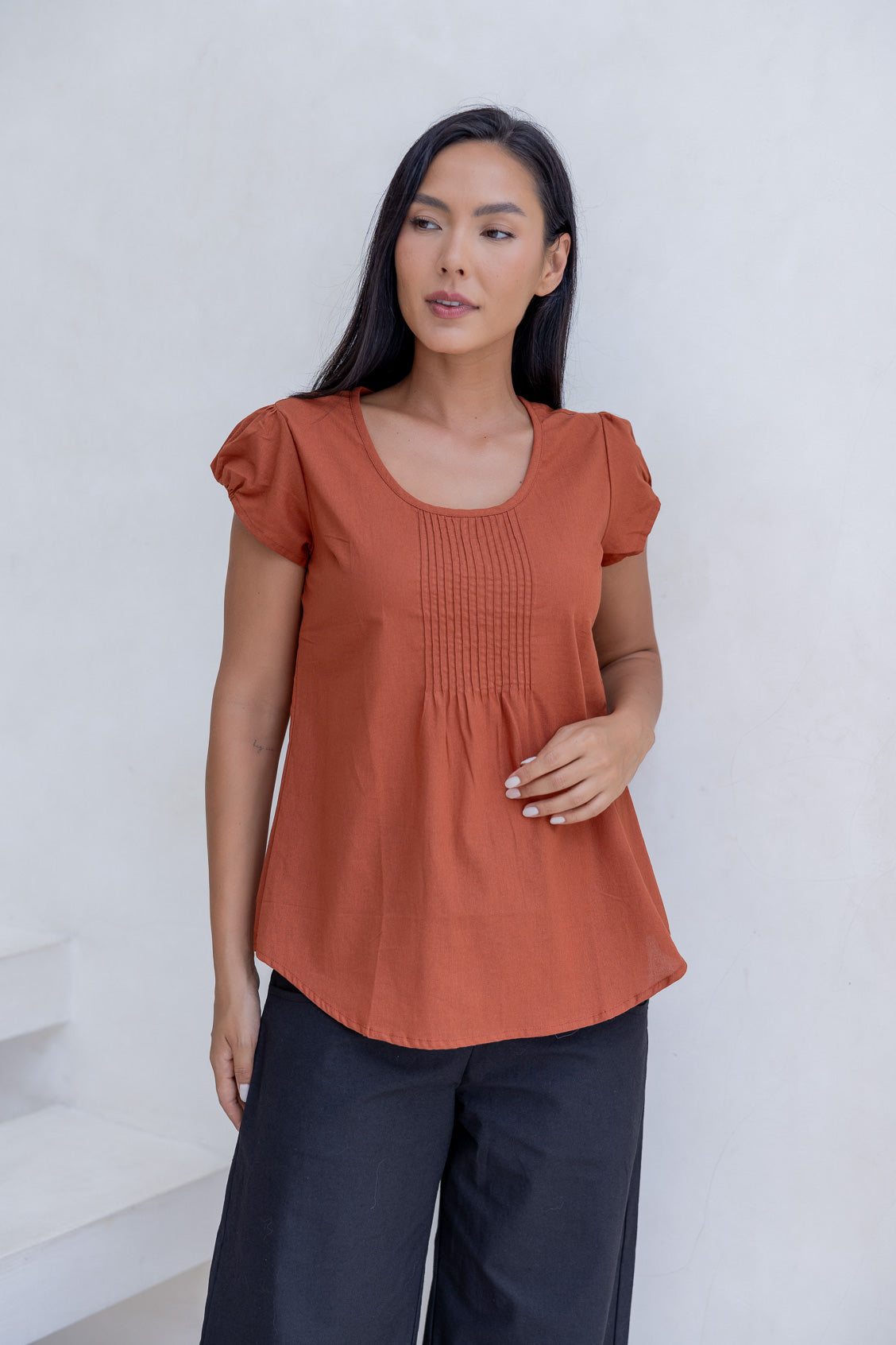 Woman wears 100% cotton cap sleeve top  in rust orange shade with pin tuck detail across the bust and a scoop neckline.
