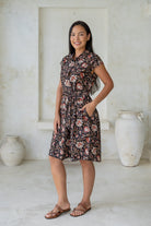 Model posing with one hand in pocket wears 100% cotton indian floral print shirt style dress with cap sleeves, fabric waist tie and box pleats at the waist.