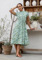 Woman wears 100% cotton button through midi length dress with short sleeves and pockets in floral print on a sage green base.