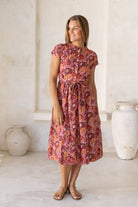 Woman poses in retro style cotton button through dress with short sleeves, high round neckline and a fabric waist tie in a warm toned floral print.