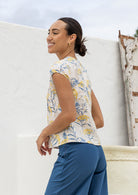 Woman stands sideways wearing a neutral based floral print cotton top standing against a white wall.