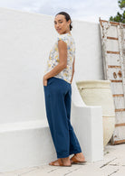 Woman stands sideways wearing a neutral based floral print cotton top standing against a white wall.
