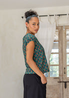 Woman stands sideways wearing a green based floral print cotton top in an indoor setting.