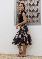 Woman standing sideways wearing a black top and black floral cotton skirt against a wooden window,