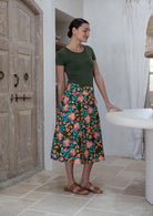 Woman standing sideways wearing a basic top and olive green based floral cotton skirt in a home setting