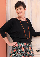 Woman wearing a 3/4 sleeve rayon batwing round neckline black top with a floral skirt and orange beads.