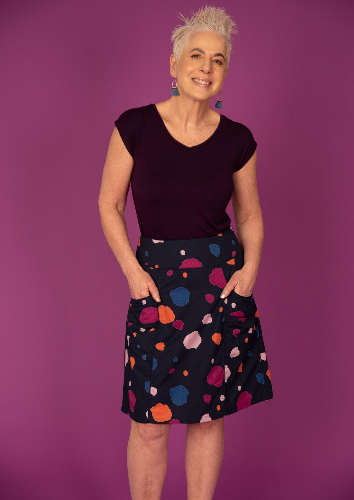 A woman standing against a purple background, wearing a knee-length skirt with a multicoloured spot print, with her hands in pockets