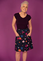 A woman standing against a purple background, wearing a knee-length skirt with a multicoloured spot print, with her hands in pockets