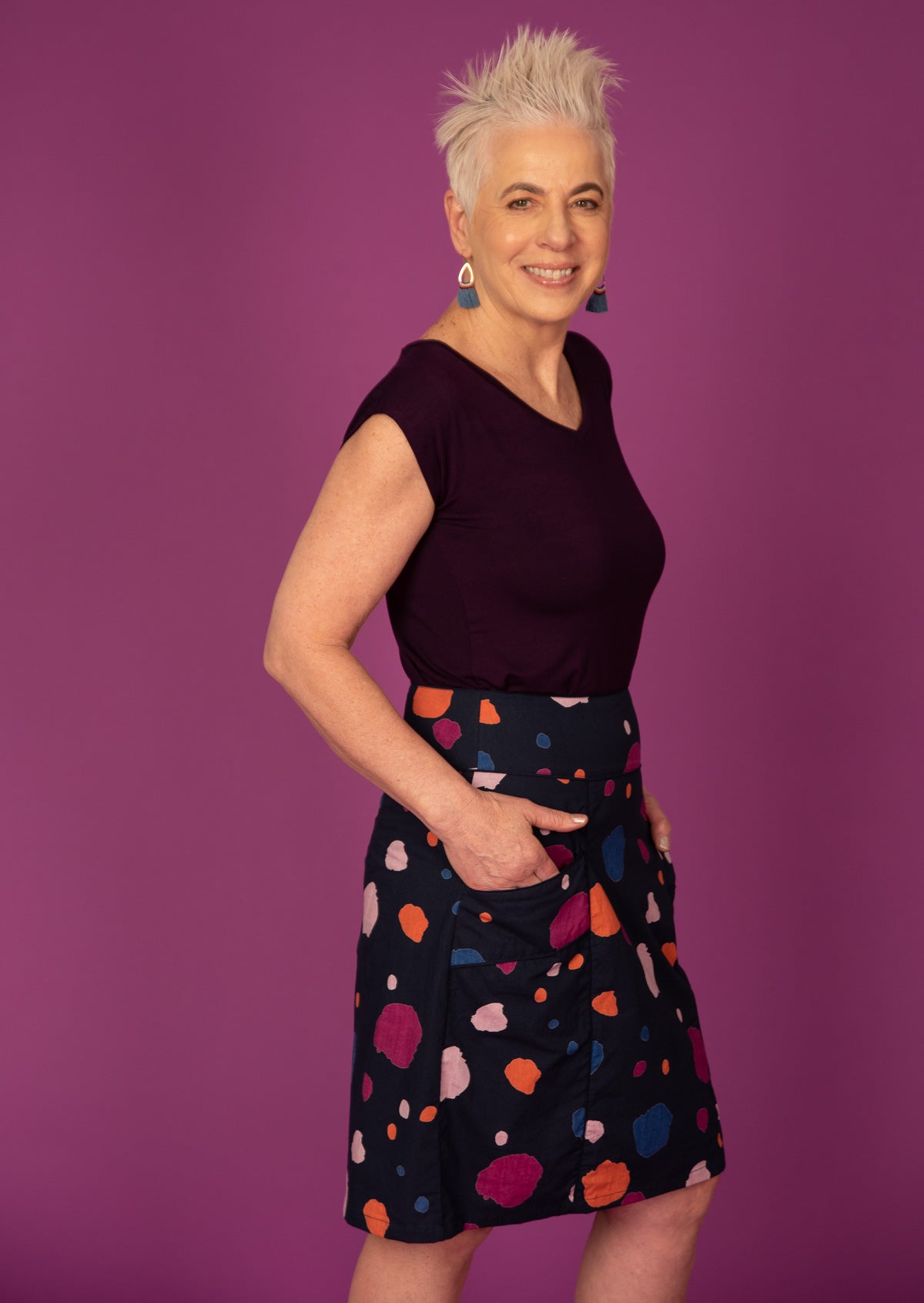 Woman stands side on in cotton knee length skirt in multi-coloured spot print on dark blue base, with hands in pockets