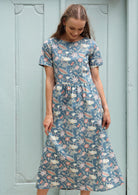 Woman stands in a short sleeve cotton calf length dress in a floral print with a mid blue base