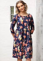 Woman wears wide round neck, 3/4 sleeve, knee length dress with pink and green toned native botanic print