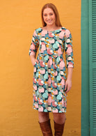 Model poses in cotton pastel print knee length dress with hand in pocket against a yellow wall