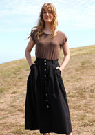 Woman with red hair wearing Cora Skirt full front button down front belt loops box pleats maxi skirt with side pockets 100% cotton black with light brown t-shirt 