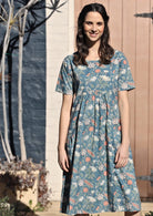 Woman wears over the knee length cotton dress with coral, sage and lemon florals on a mid blue base